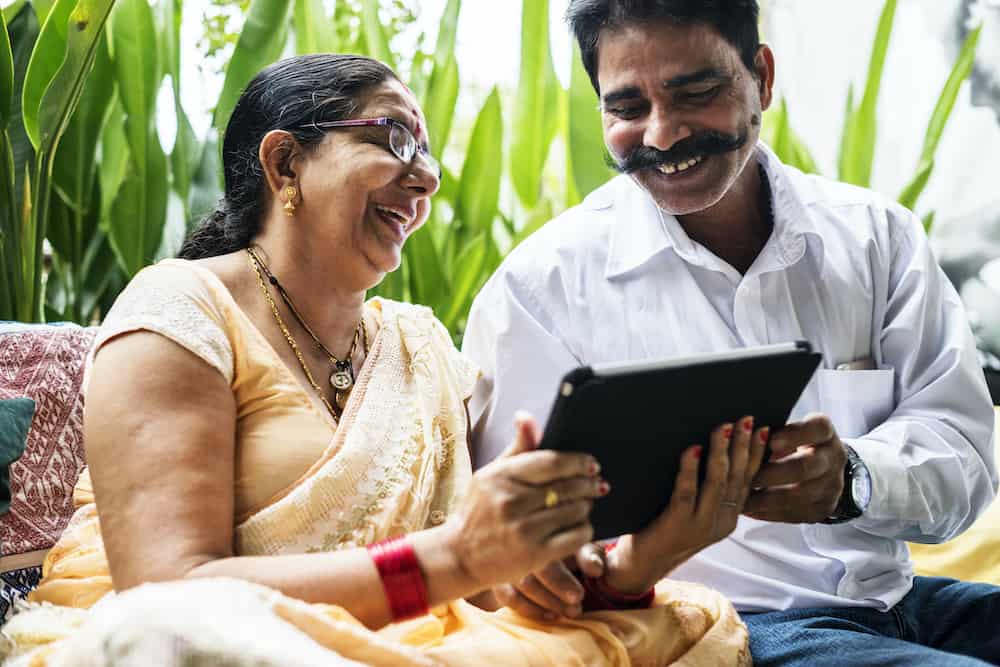 Joyful middle-aged Indian couple sharing a laugh while looking at a tablet, sitting in an indoor setting with green foliage, symbolizing happiness and shared experiences.