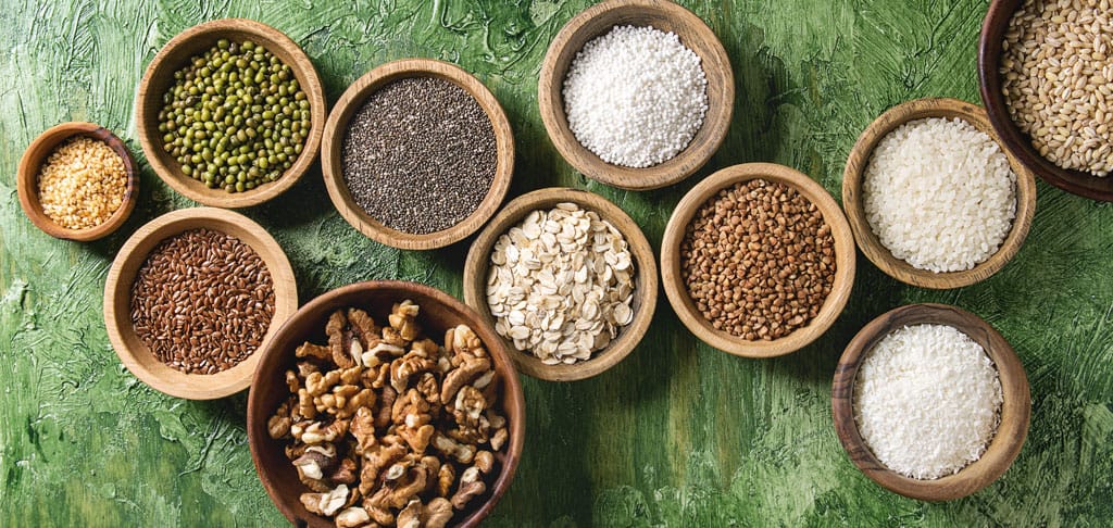 Collection of wooden bowls filled with various grains, seeds, and nuts including mung beans, lentils, chia seeds, flaxseeds, oat flakes, buckwheat, walnuts, sesame seeds, and rice, arranged on a green background, symbolizing the focus on healthful, whole foods in the 'About Us' page.