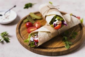 Freshly prepared wrap cut in half and wrapped in parchment paper, served on a round wooden board with pickles, cherry tomatoes, and fresh herbs, accompanied by a bowl of dipping sauce.