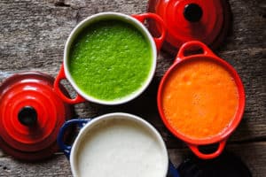 Three colorful stoneware pots containing green, white, and orange soups, possibly spinach, cream-based, and carrot soups respectively, arranged on a rustic wooden surface, symbolizing the variety of healthy soups featured in the 'Daal-Palak Ka Shorba (Lentil and Spinach Soup)' page.