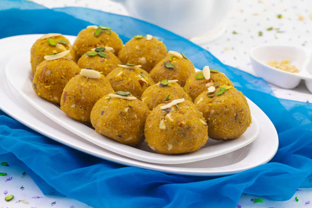 Image of a plate of golden-brown Indian sweets known as 'laddoos', garnished with slivers of almonds and pistachios, placed on a white surface with a vibrant blue cloth underneath, as featured on the 'Methi Ladoo' page.