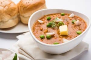 Image of a bowl of rich, creamy curry garnished with butter, peas, onions, and cilantro, served with fluffy bread rolls, as featured on the 'Pav Bhaji' page.