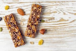 Image of three nutritious granola bars densely packed with nuts, seeds, and dried fruits, drizzled with chocolate, and surrounded by individual ingredients, as featured on the 'Dry Fruit Protein Bar' page.