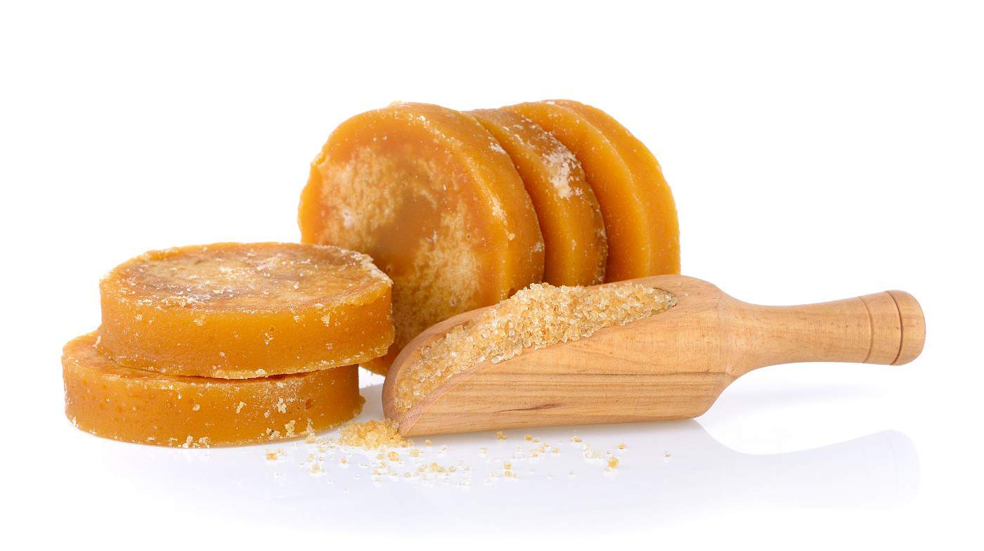 Image of several pieces of palm sugar stacked with granulated sugar spilling from a wooden scoop, highlighting the coarse and granular texture of the sugar, as featured on the 'The Benefits of Organic jaggery' page.