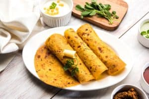 Image of three spiced Indian flatbreads with a pat of melting butter, served with a yogurt-based dip and fresh herbs, accompanied by green and red sauces, as featured on the 'Methi Thepla' page.