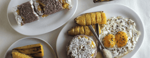 A plate of fried eggs and sliced plantains, a dish of white seasoning, and two plates of pastries possibly rolled in coconut flakes with a dark filling, suggesting a breakfast or brunch with a tropical or Latin American cuisine influence.