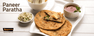 A plate of Paneer Paratha, accompanied by a bowl of chutney and bowls of paneer, set on a white textured surface.